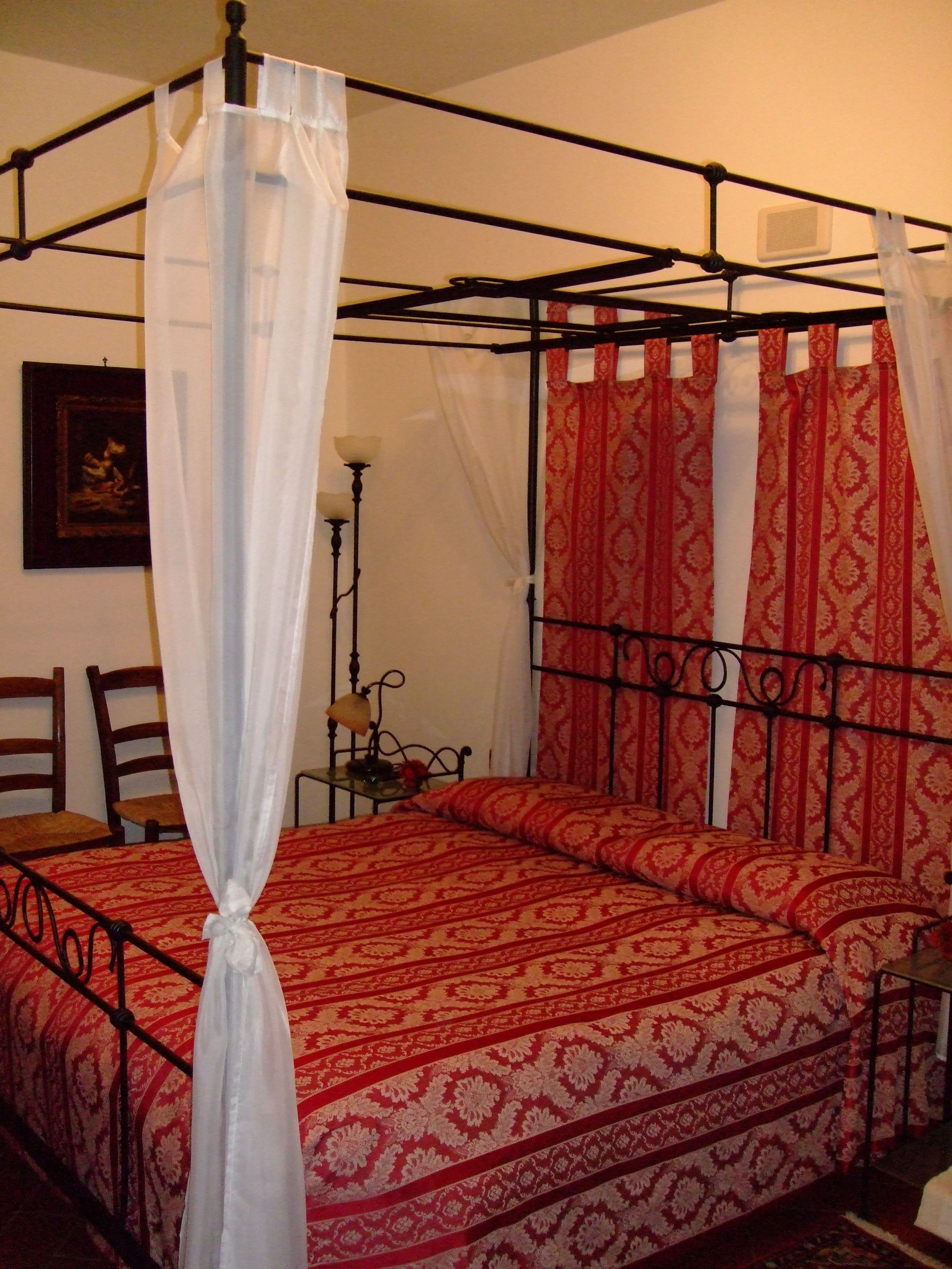 A four-poster bed with a canopy, red patterned bedding, and a small bedside table with a lamp.
