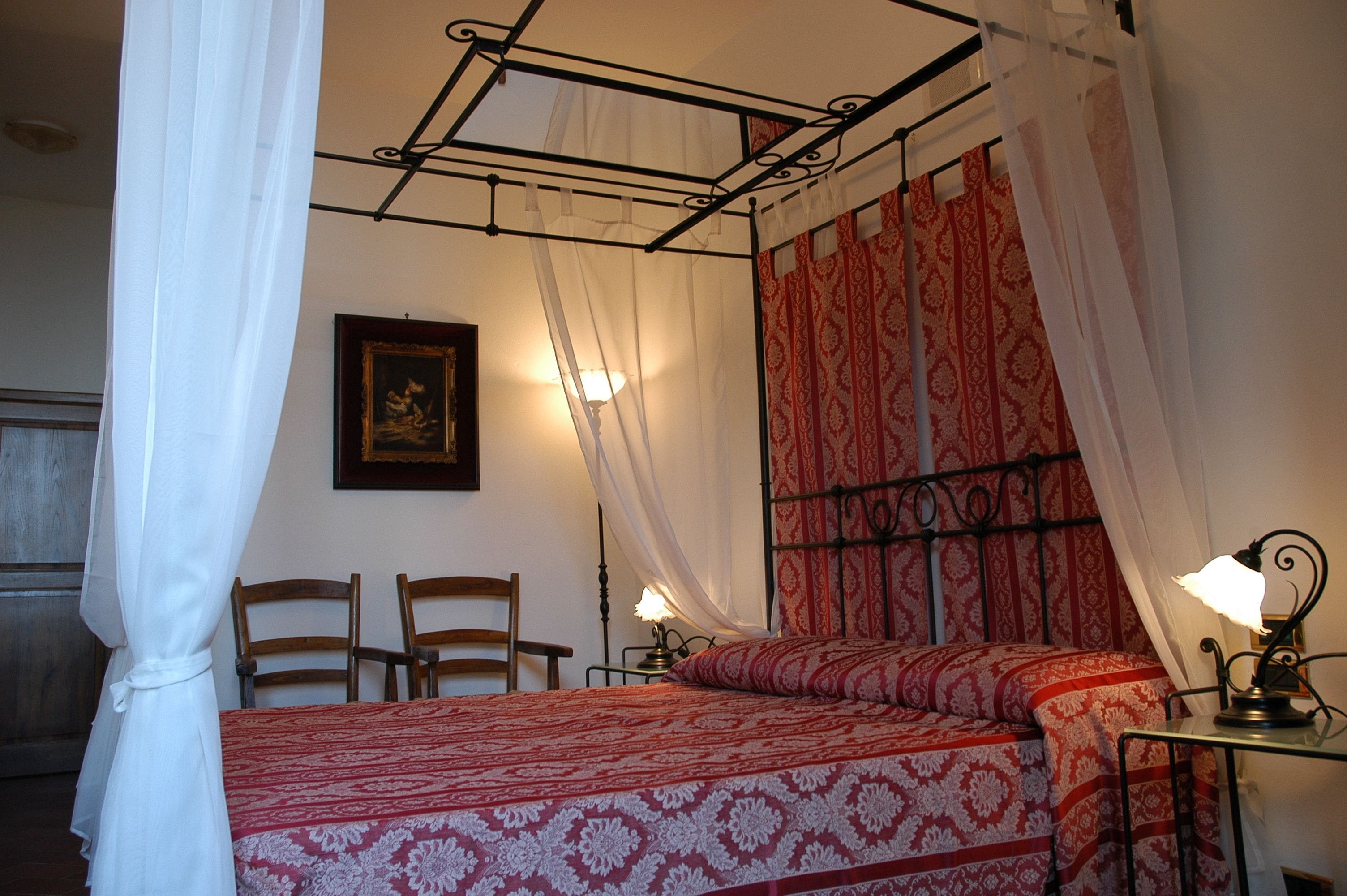 Ein Schlafzimmer mit Himmelbett, Nachttisch mit Lampe, zwei Stühlen und einem gerahmten Bild an der Wand.