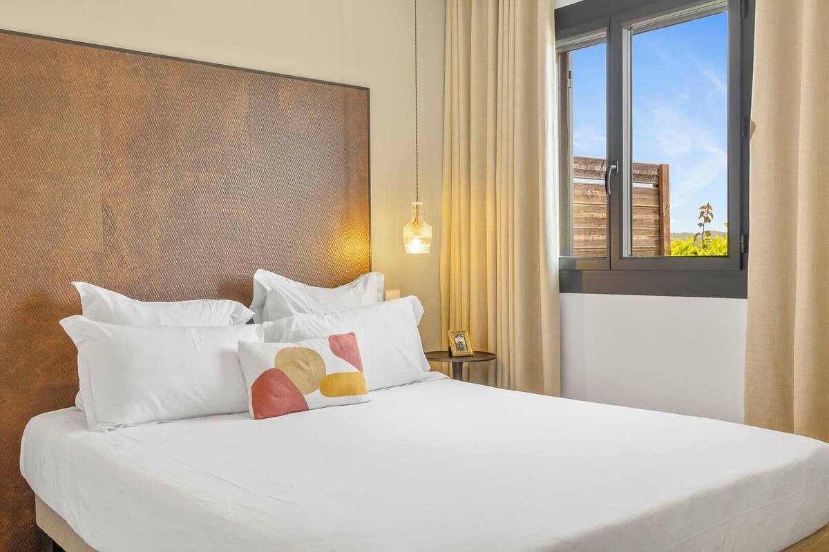 A hotel room with a large bed, a headboard, a window with curtains, and a small table with a lamp.
