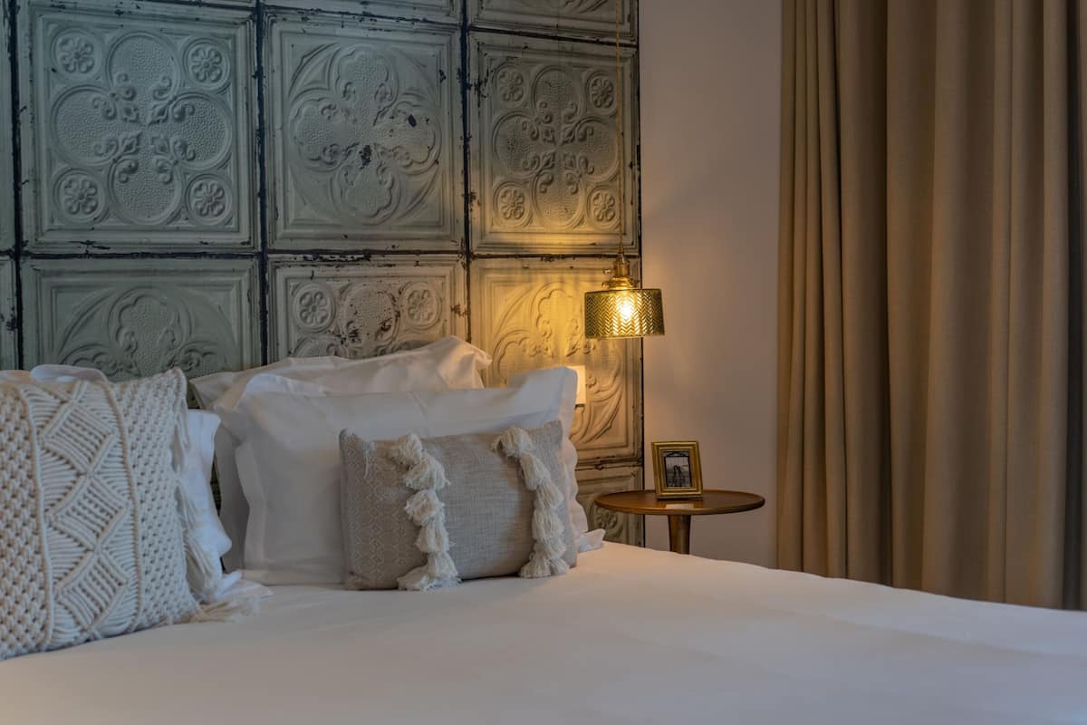 A neatly made bed with white linens and decorative pillows, a wall with intricate paneling, a small round table with a framed picture, and a lamp with a gold shade.