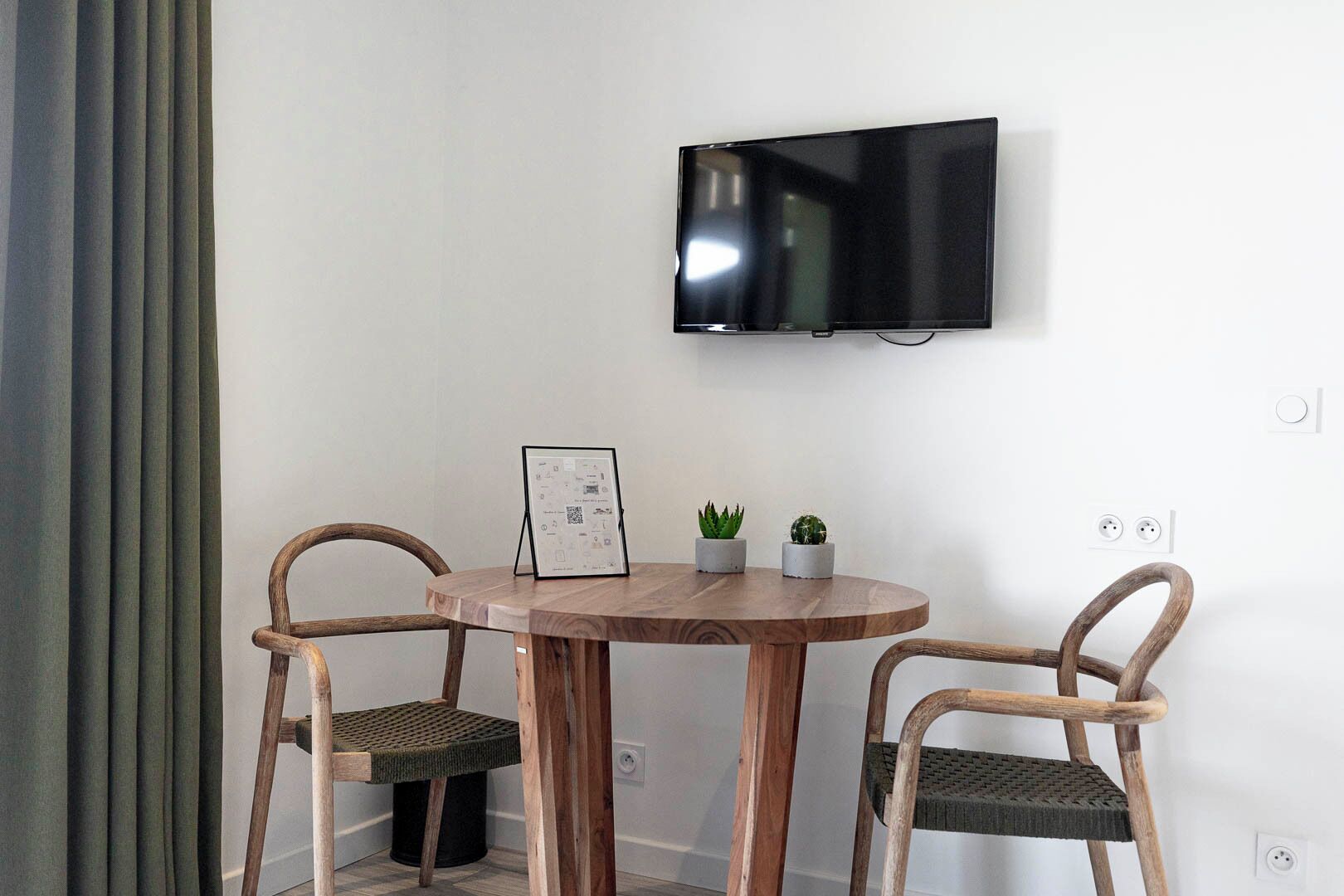 Une petite pièce avec une table ronde en bois, deux chaises en osier, un tableau encadré, deux petites plantes en pot et un téléviseur fixé au mur.