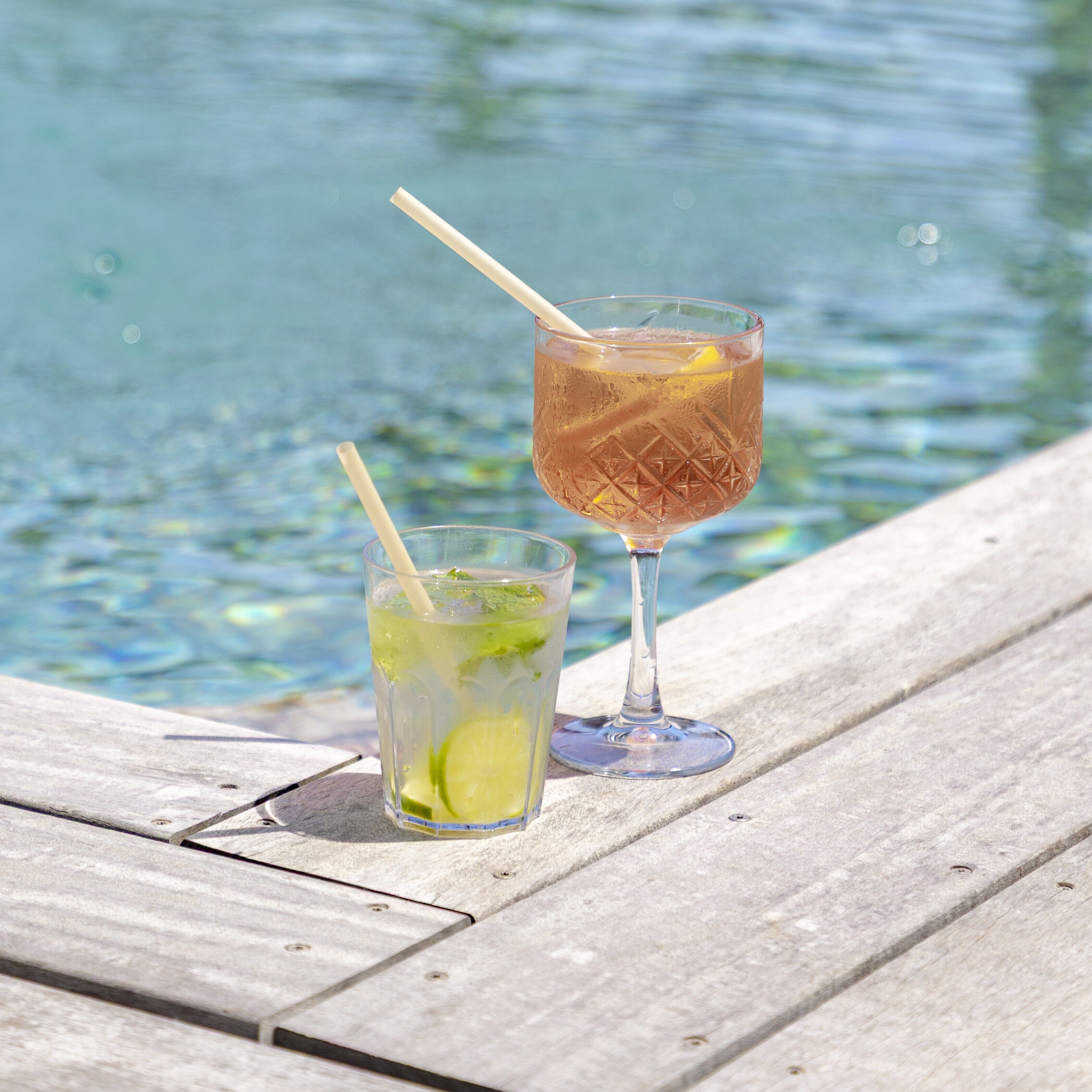 Two glasses of a beverage with lime and mint on a wooden dock by a body of water.