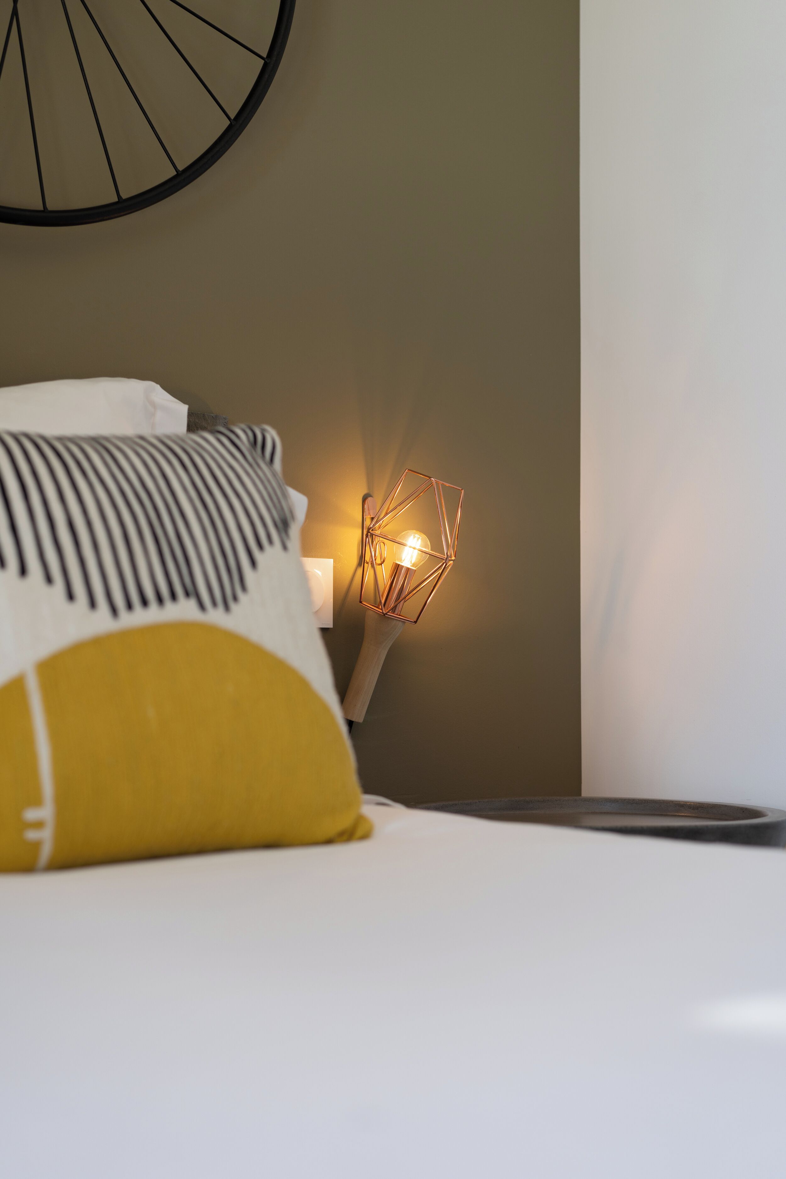 Una cama bien hecha con un cojín blanco y amarillo, una lámpara de noche con diseño geométrico y un reloj de pared.