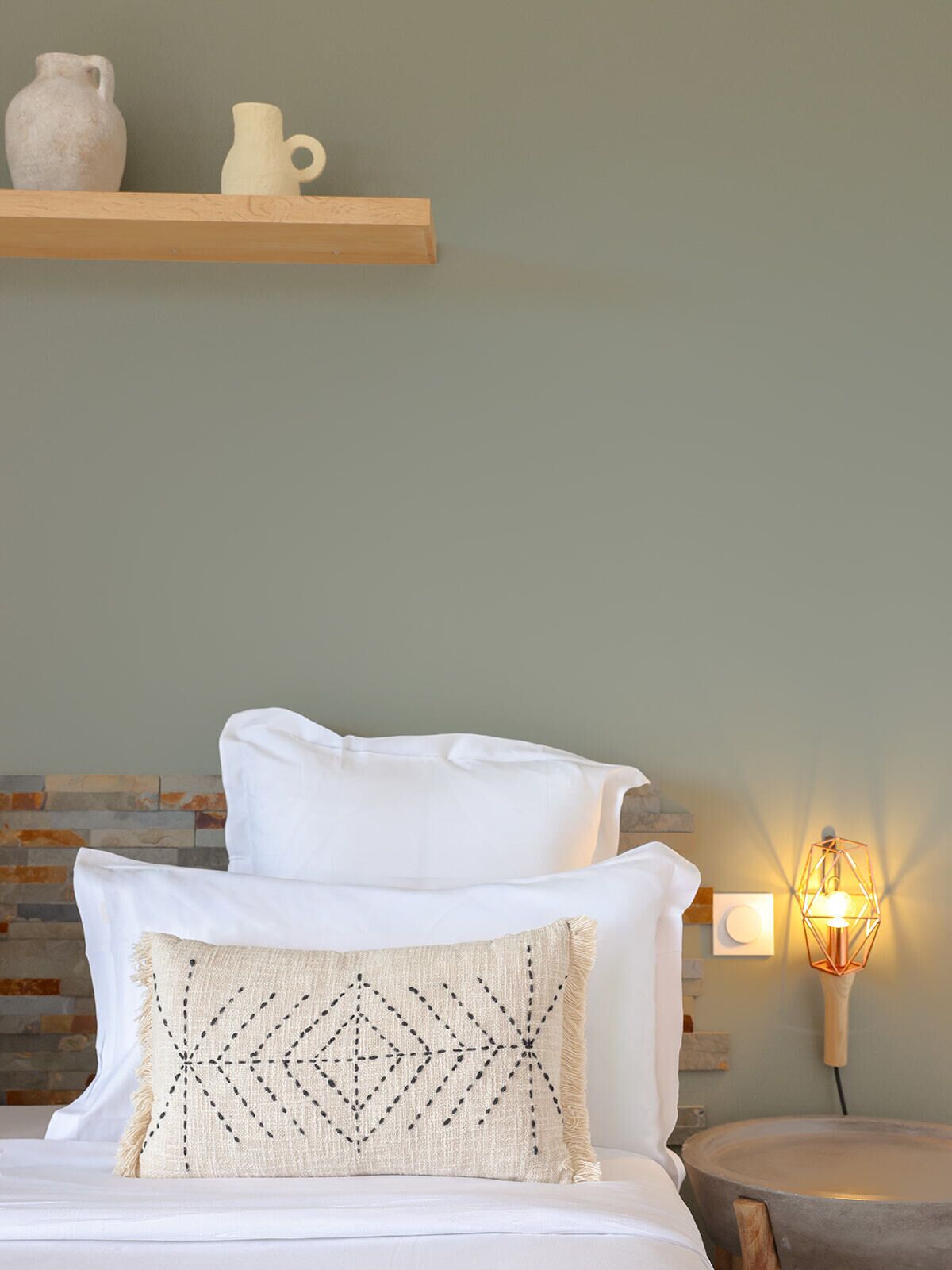 A bedroom with a bed, pillows, a nightstand, a wall-mounted lamp, and a shelf with two ceramic pitchers.