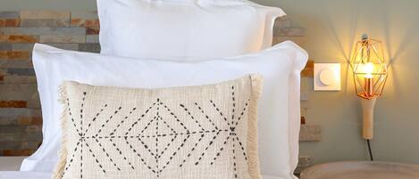 A bedroom with a bed, pillows, a nightstand, a wall-mounted lamp, and a shelf with two ceramic pitchers.