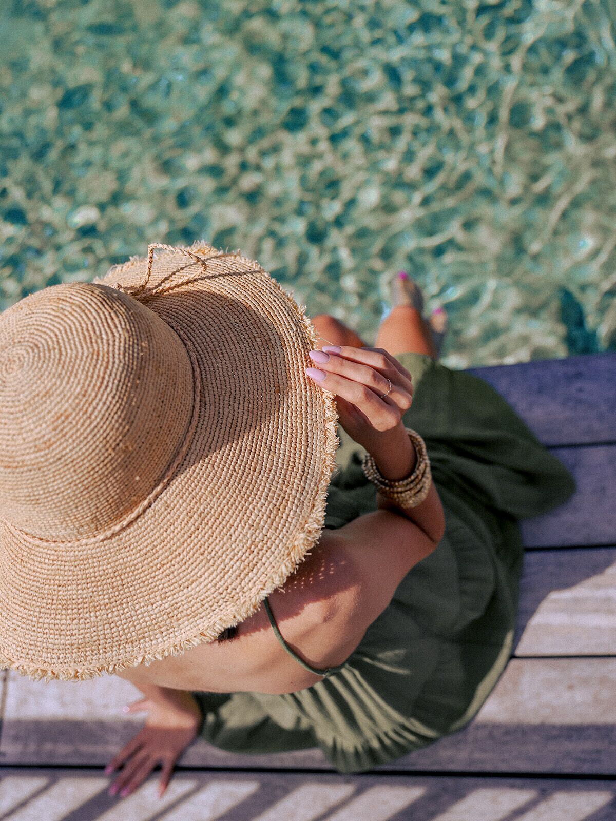 A person sitting on a wooden deck with a woven hat, wearing a green dress, and holding a small object.