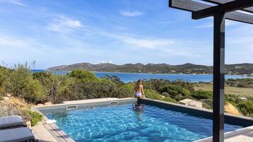 A pool with clear blue water, surrounded by wooden deck and lounge chairs, overlooking a scenic coastal landscape.