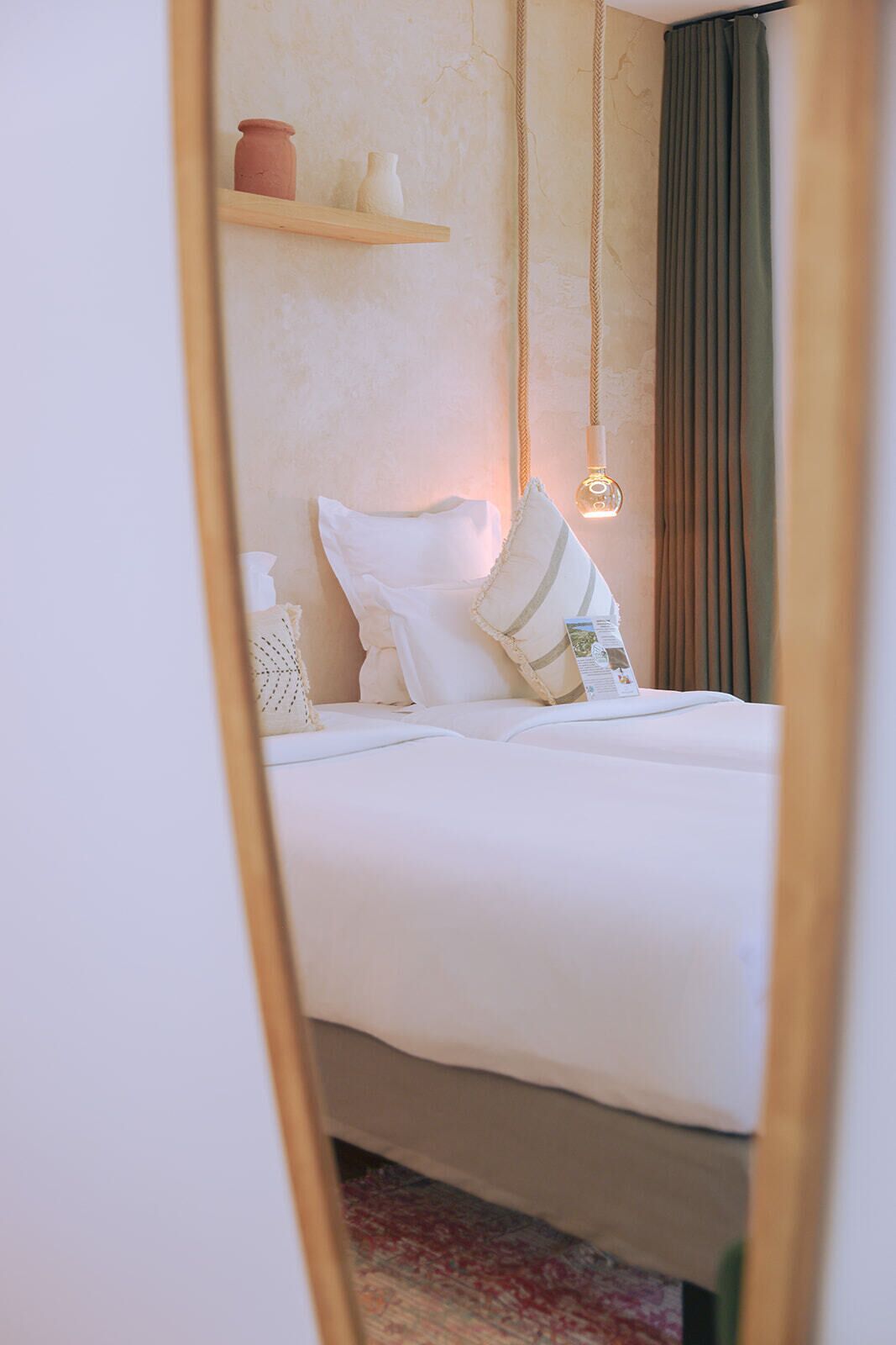 Una cama con ropa blanca, un colgante de luz y un estante con adornos.