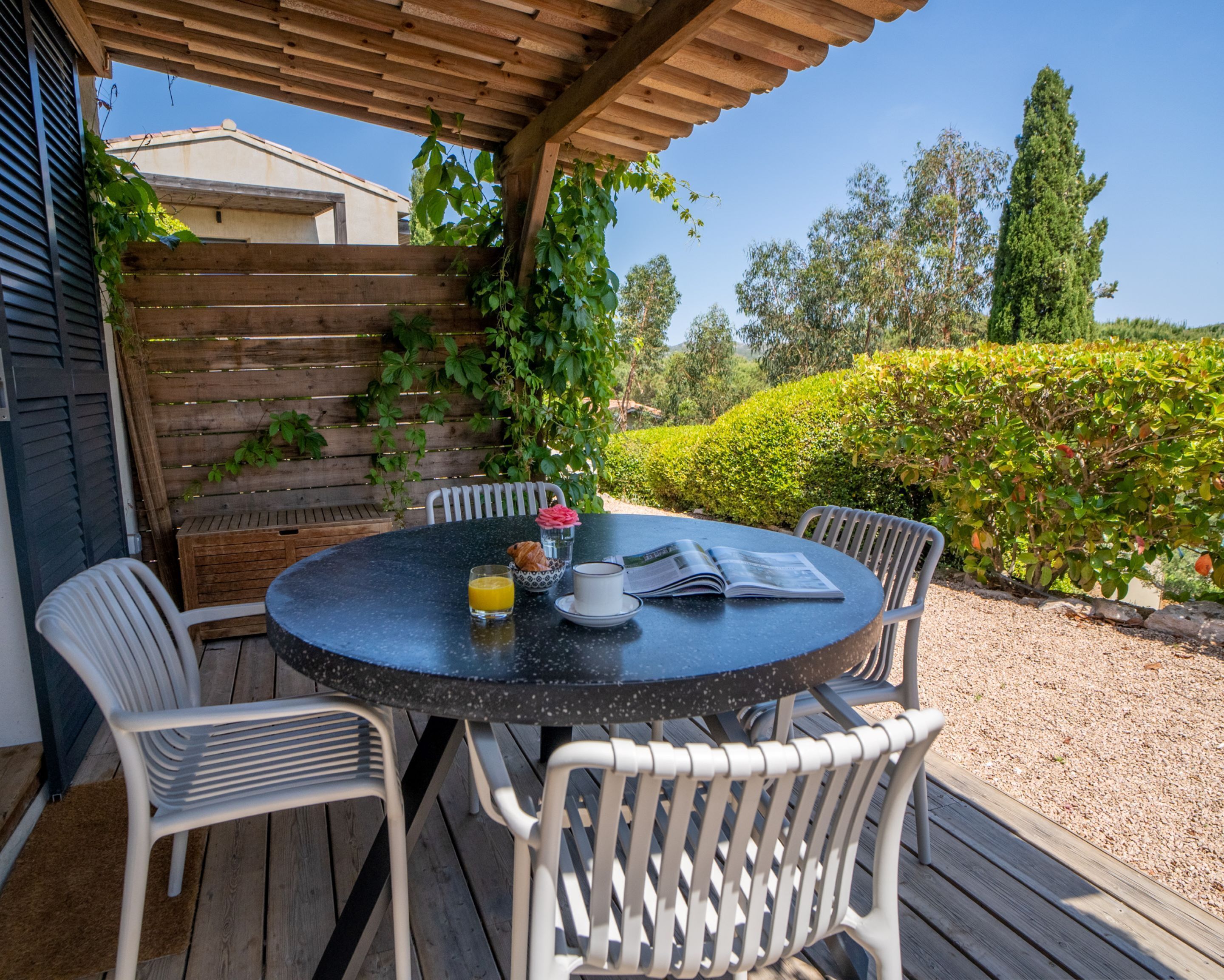 Ein runder Tisch mit vier weißen Stühlen, eine hölzerne Pergola darüber und Blick auf einen Garten mit Hecke und Bäumen.