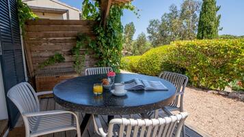 Une table ronde entourée de quatre chaises blanches, une pergola en bois au-dessus, et une vue sur un jardin agrémenté d’une haie et d’arbres.
