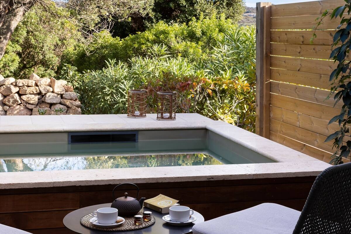 A patio area with a hot tub, two chairs, a table with a teapot and cups, and a view of a garden and trees.