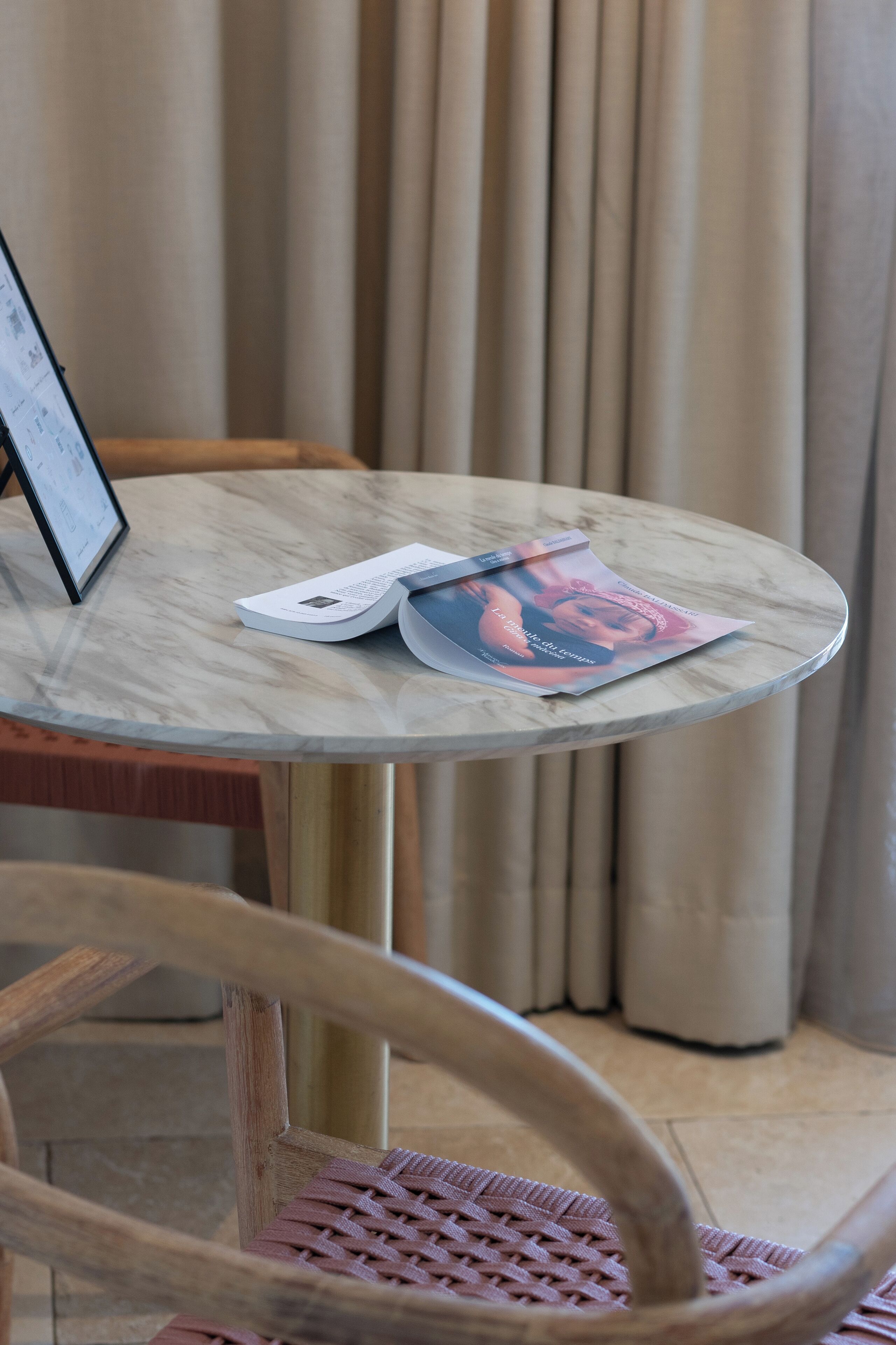 Una mesa redonda de mármol con una revista y un menú encima, una silla de madera en primer plano y una cortina al fondo.