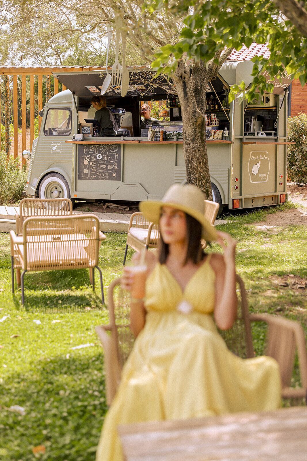 A vintage truck with a food service setup, a woman in a yellow dress sitting on a wicker chair, and a tree in the background.