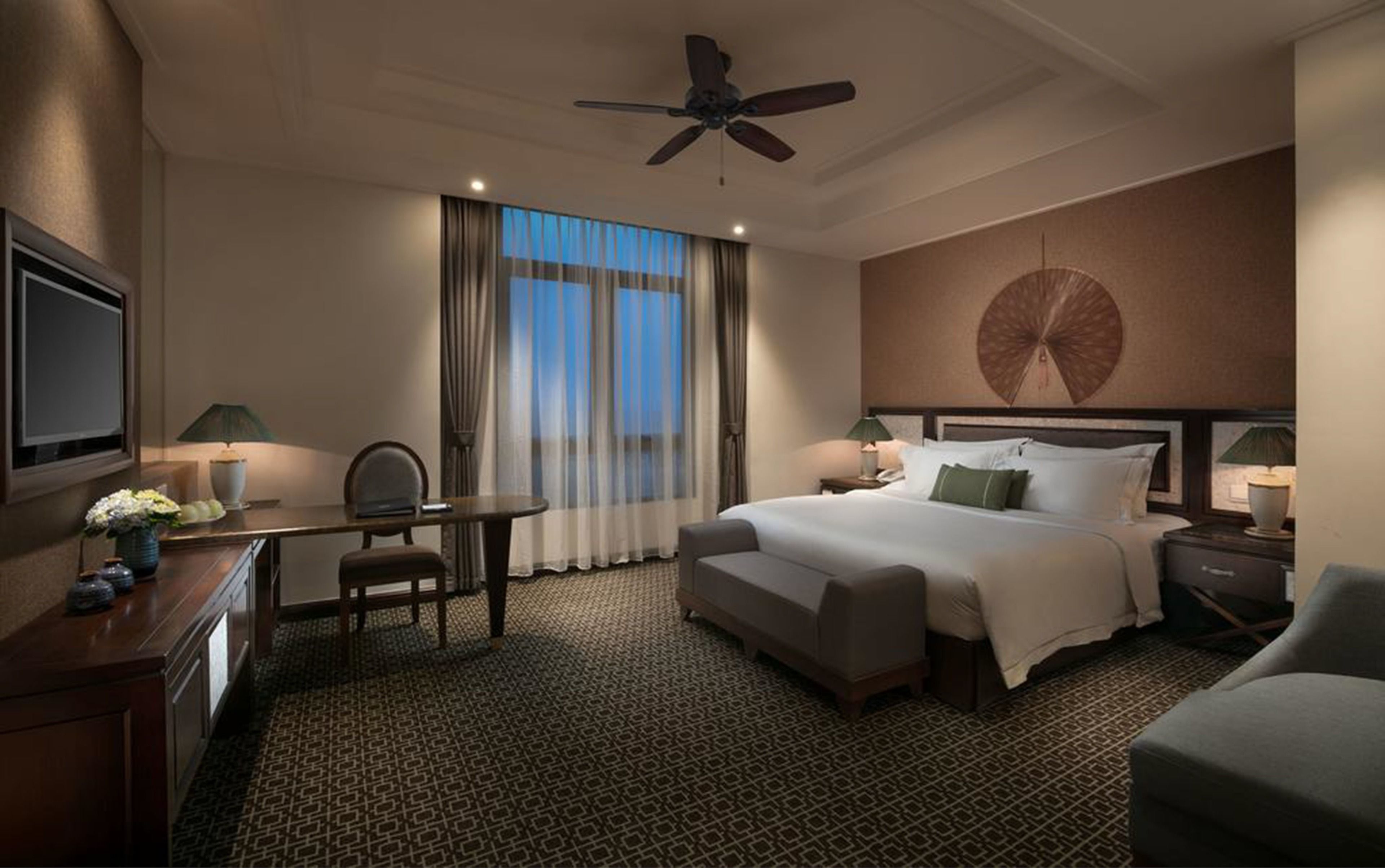 Ein Hotelzimmer mit einem großen Bett, einem Schreibtisch, einem Sessel, einem Fernseher und einem Deckenventilator.