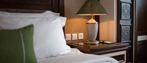 A neatly made bed with white linens and a green pillow, a bedside table with a lamp, and a wall with a textured pattern and a power outlet.