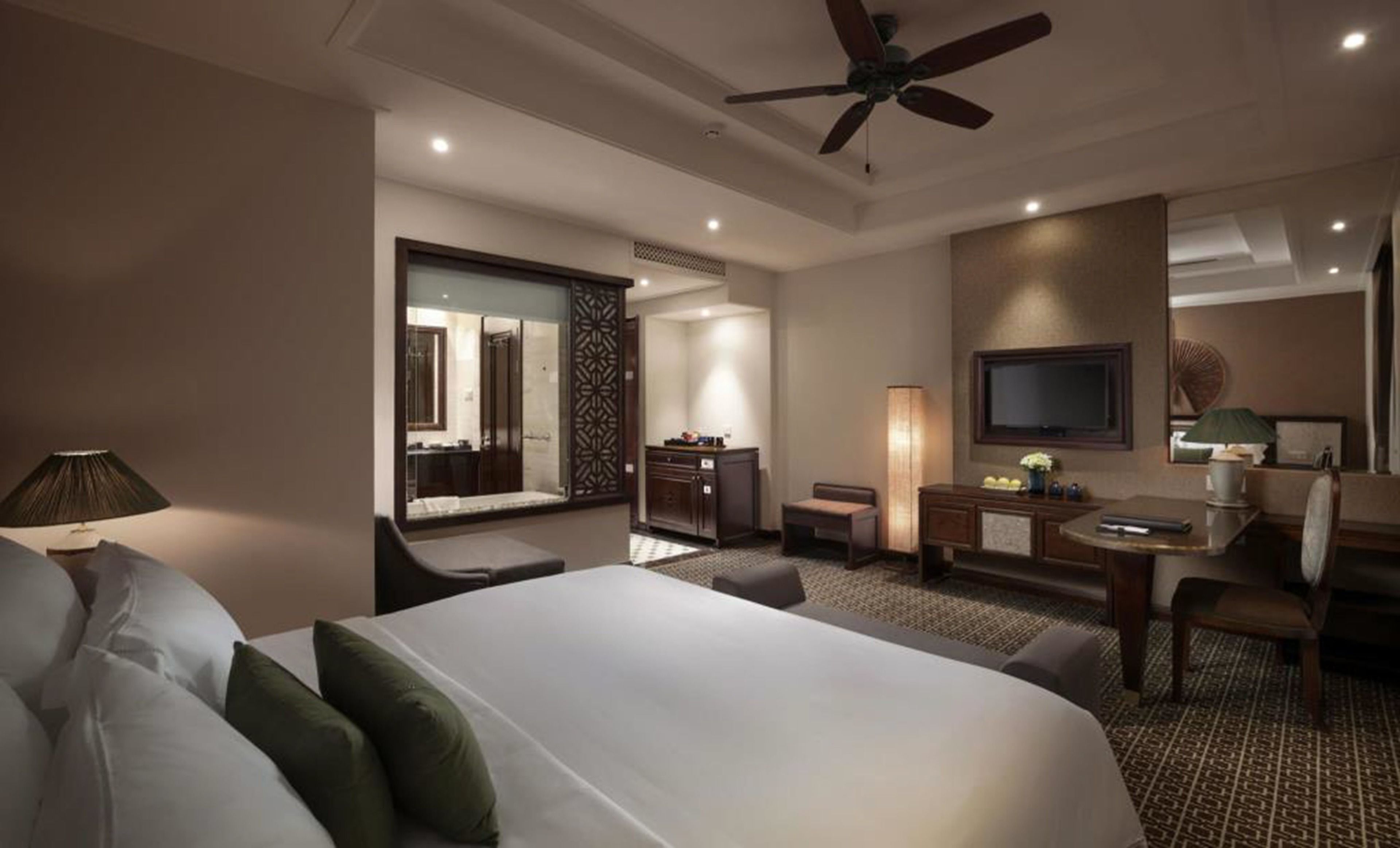 A hotel room with a large bed, a desk, a chair, a TV, and a ceiling fan.