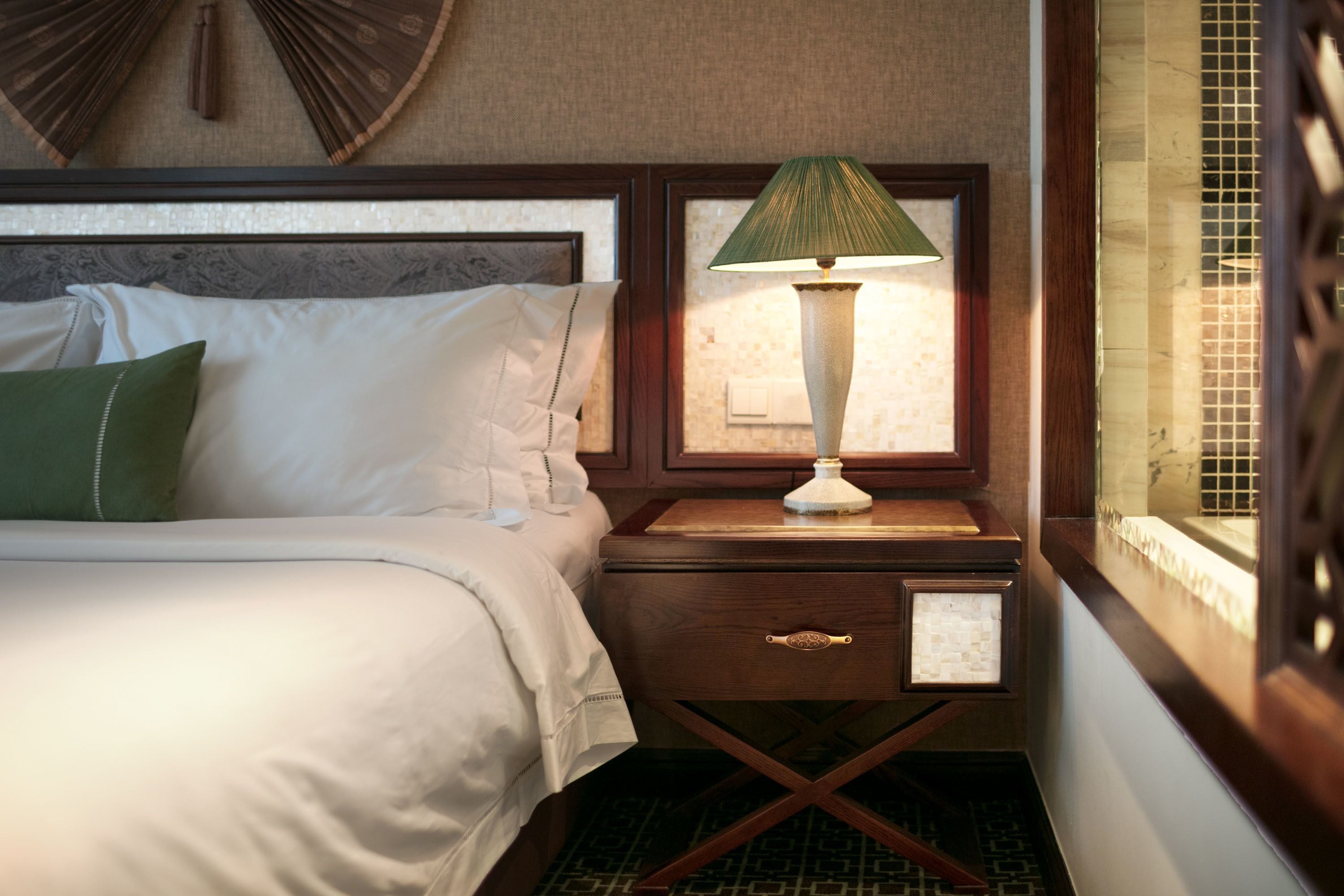 A neatly made bed with white linens and a green pillow, a bedside table with a lamp, and a window with a view of a tiled wall.