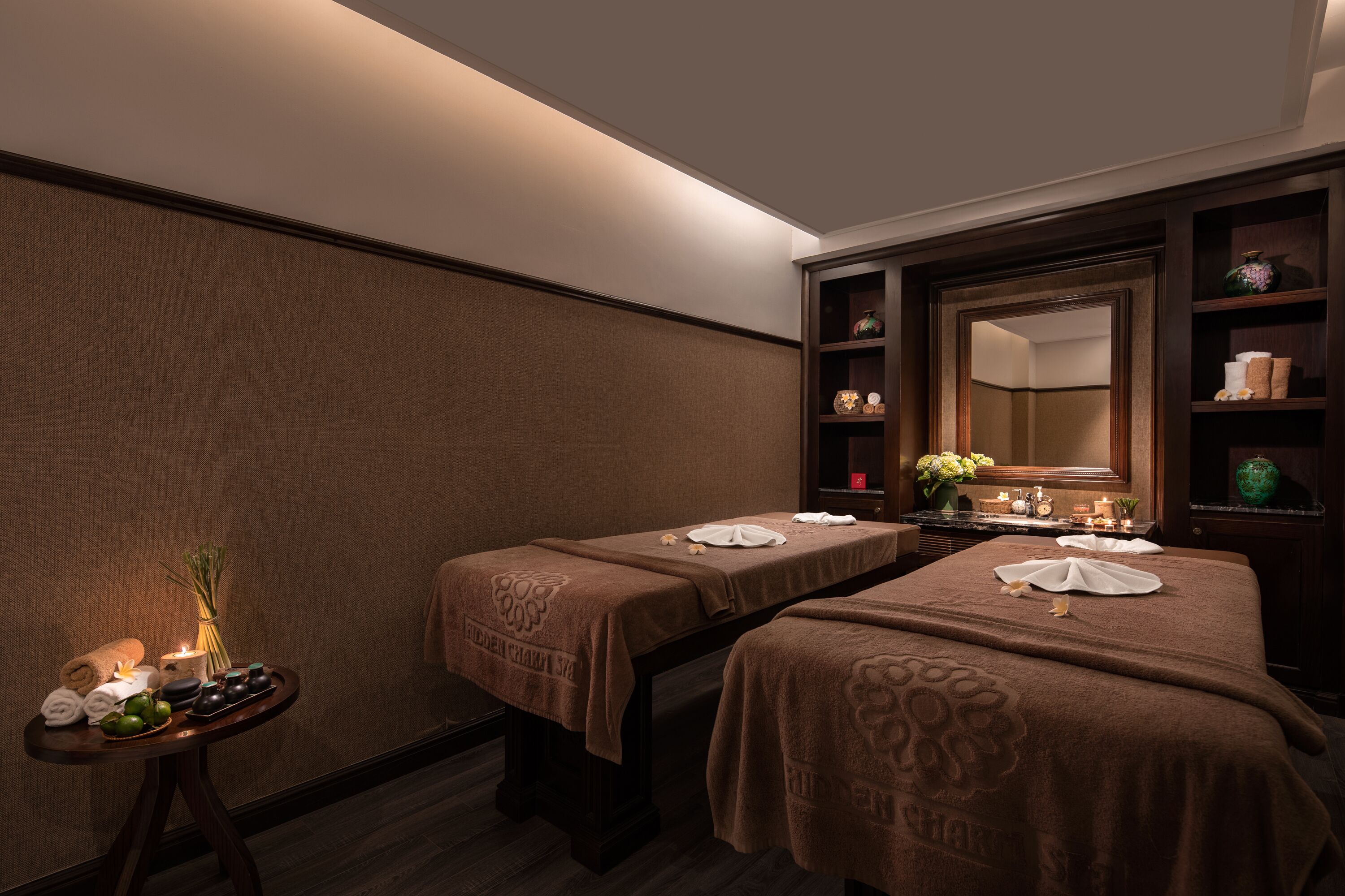 A hotel room with two beds, a small table with toiletries, a mirror, and a shelf with decorative items.
