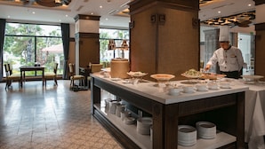 Un buffet où un chef prépare et sert les plats, un espace repas avec des chaises et une table, et une vue sur l’extérieur grâce à de grandes fenêtres.