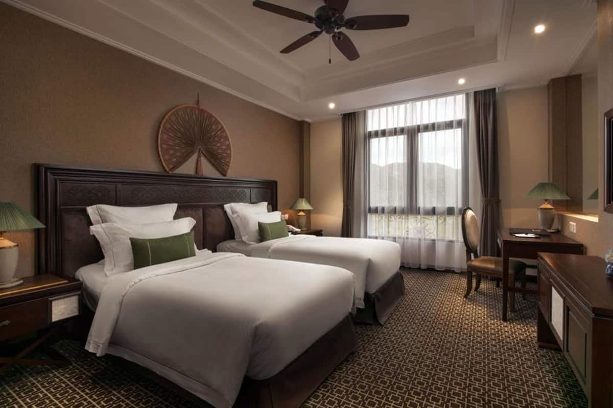 Une chambre d’hôtel avec deux lits, un ventilateur de plafond, une grande fenêtre avec des rideaux, un bureau avec une lampe et un téléviseur fixé au mur.