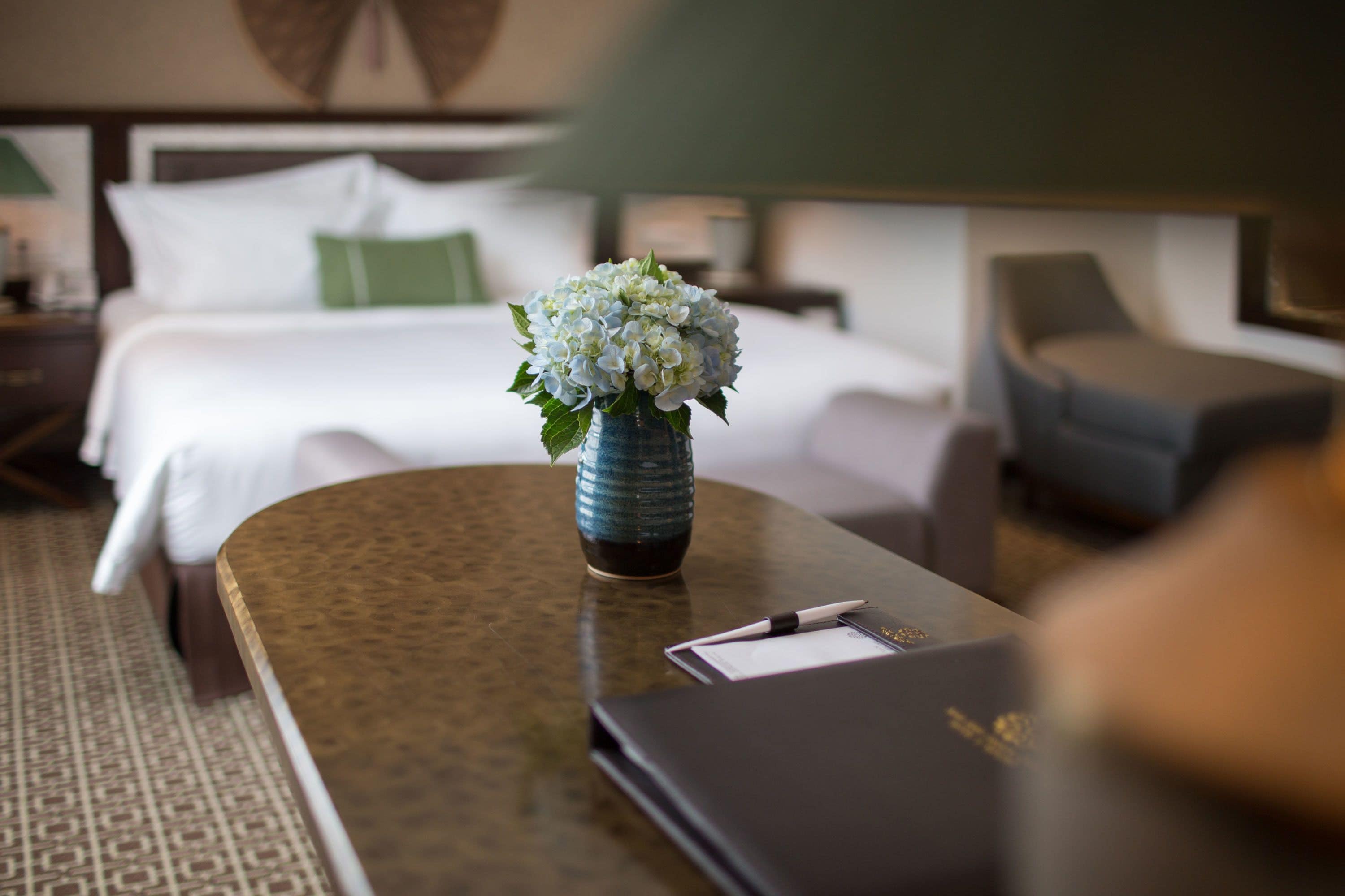 A hotel room with a neatly made bed, a bedside table with a vase of flowers, and a chair.