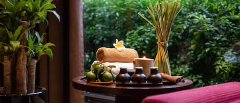 A spa setting with a table featuring a towel, candles, and small vases, surrounded by greenery.