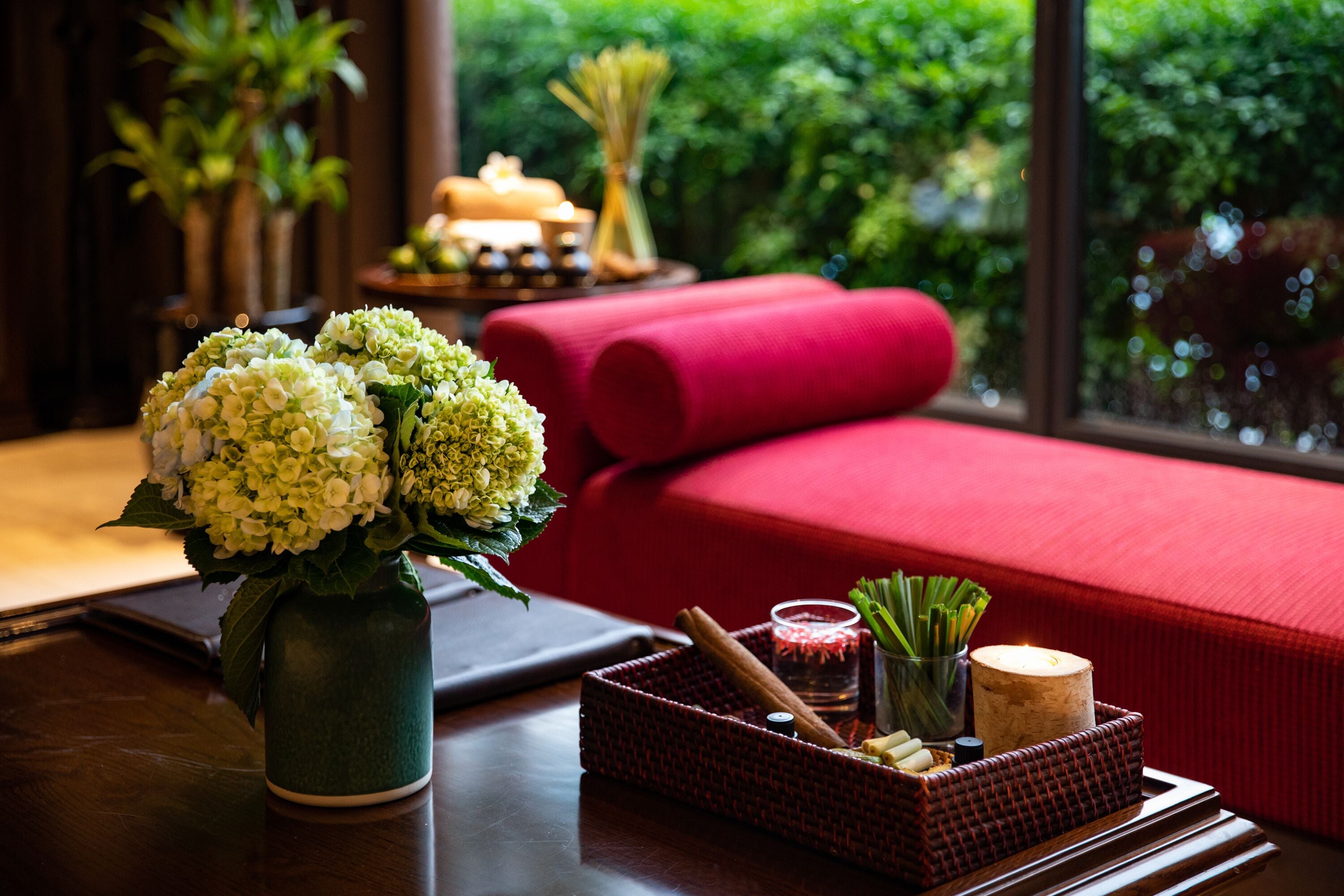 A room with a wooden table, a vase of white flowers, a red sofa, and a window with a view of greenery.