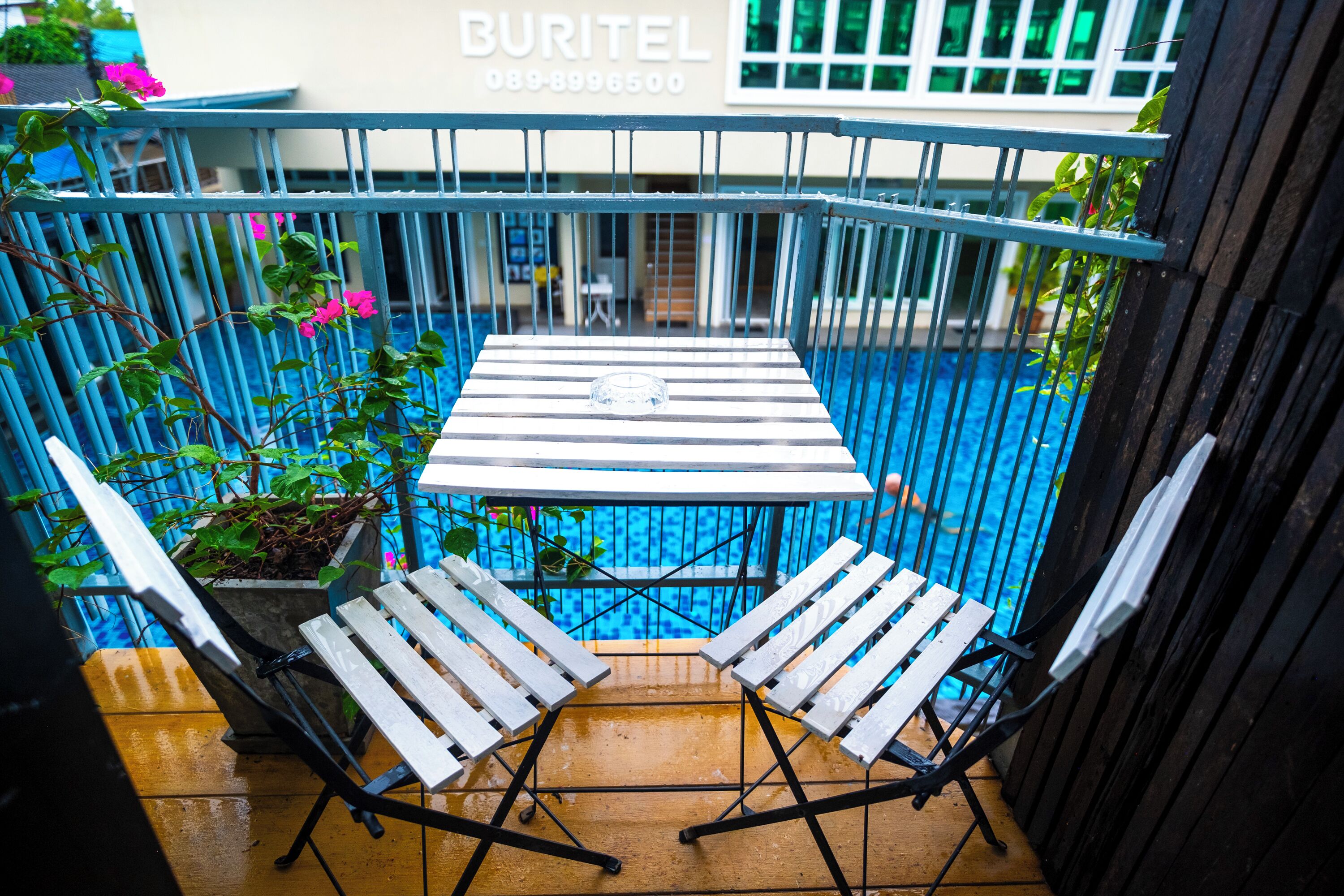 Un balcón con una mesa y dos sillas, una piscina al fondo y un edificio con un letrero.