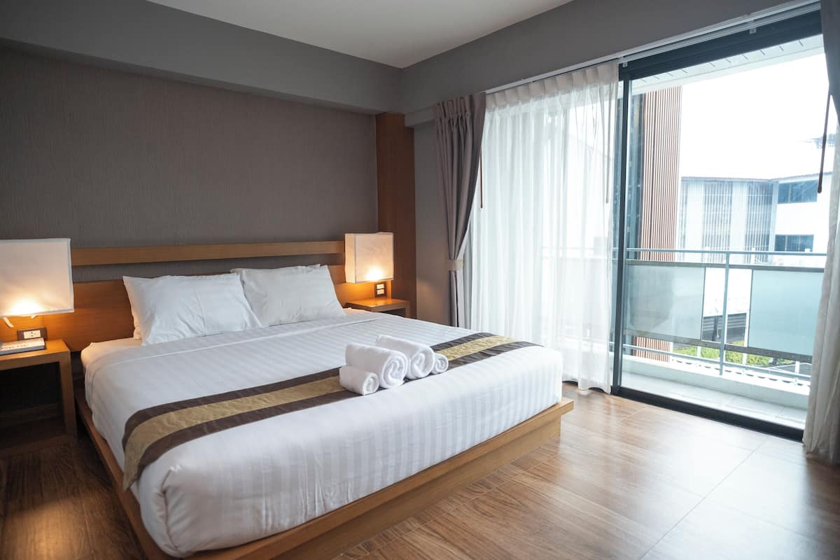 A hotel room with a large bed, two bedside tables with lamps, a view of the outside through a sliding glass door, and a balcony.