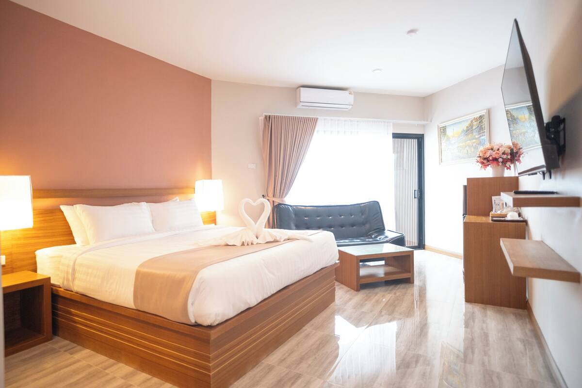 A hotel room with a large bed, bedside tables, a desk, a chair, a TV, and an air conditioning unit.