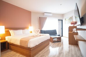 A hotel room with a large bed, bedside tables, a desk, a chair, a TV, and an air conditioning unit.