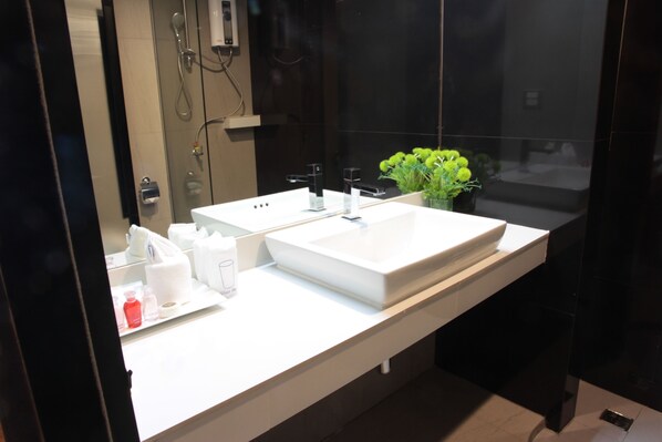 A modern bathroom with a large white sink, two faucets, and a mirror. There are toiletries on the counter and a small plant on the right.