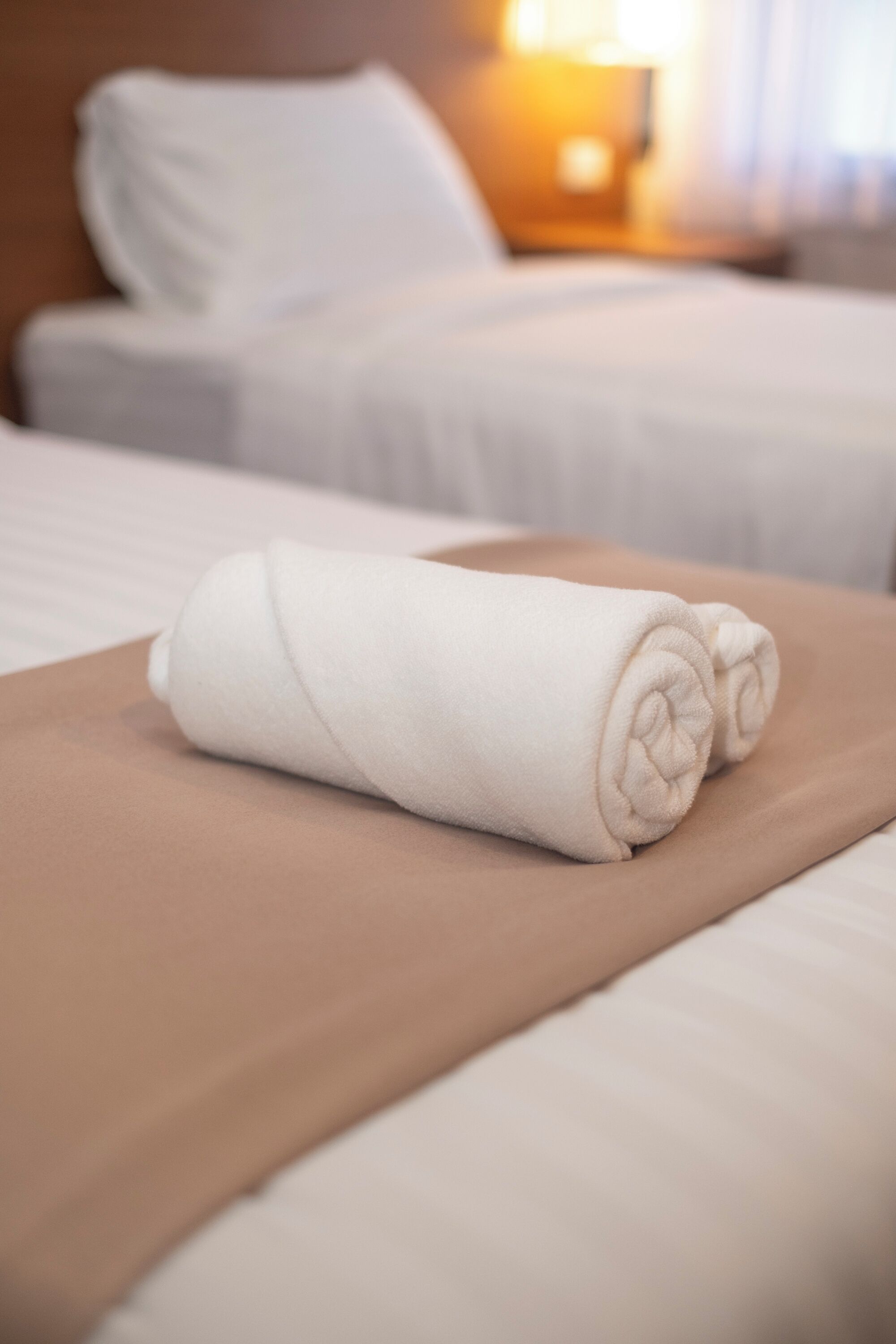 Una toalla blanca bien doblada sobre una cama con una colcha marrón, una almohada blanca y una mesita de noche con una lámpara al fondo.