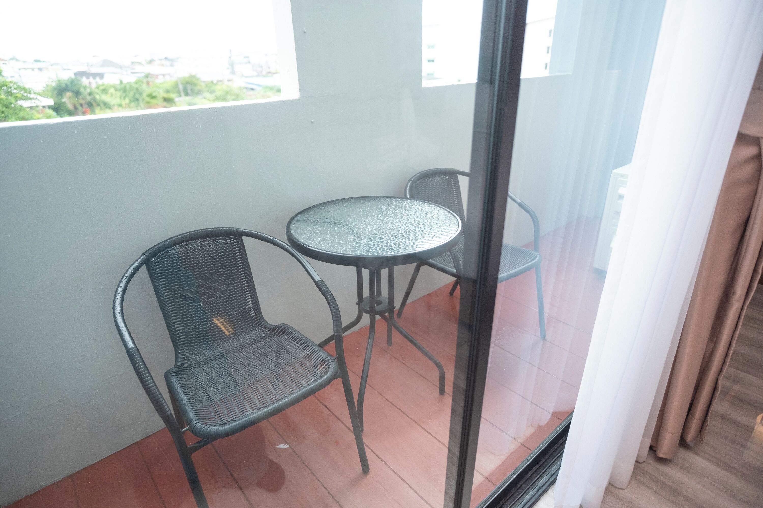Un balcón con dos sillas de metal, una mesa pequeña, una puerta de cristal y vistas a zonas verdes y edificios.