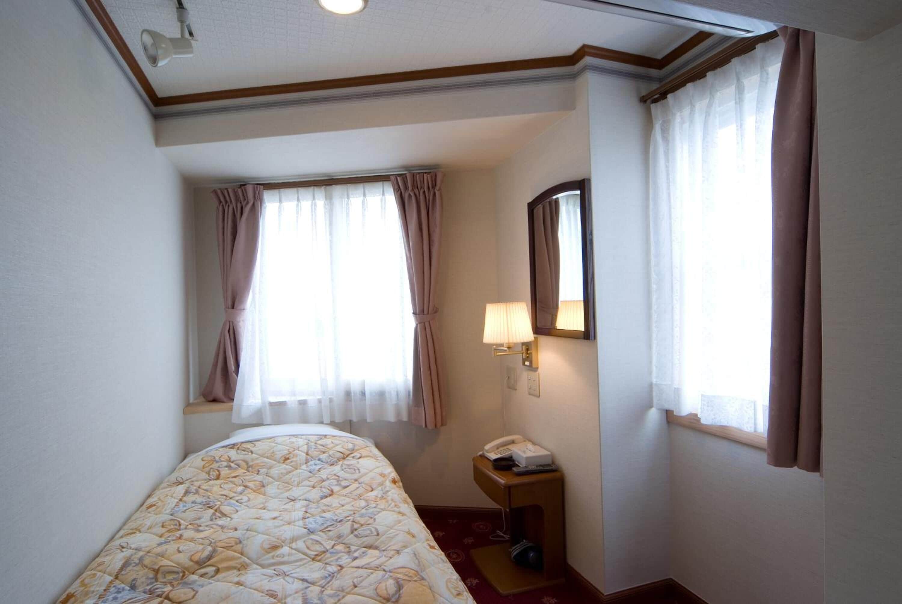 A hotel room with a bed, a bedside table with a lamp, a telephone, and a window with curtains.