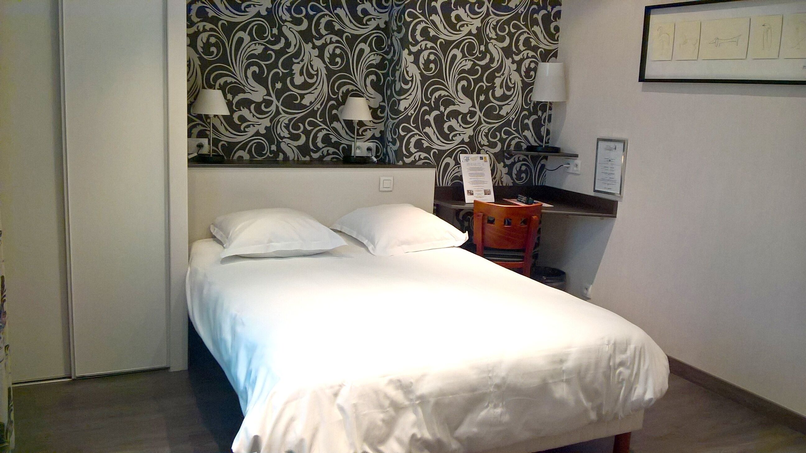 Una cama pequeña y bien hecha con sábanas blancas en una habitación con papel tapiz estampado y un escritorio con una silla.