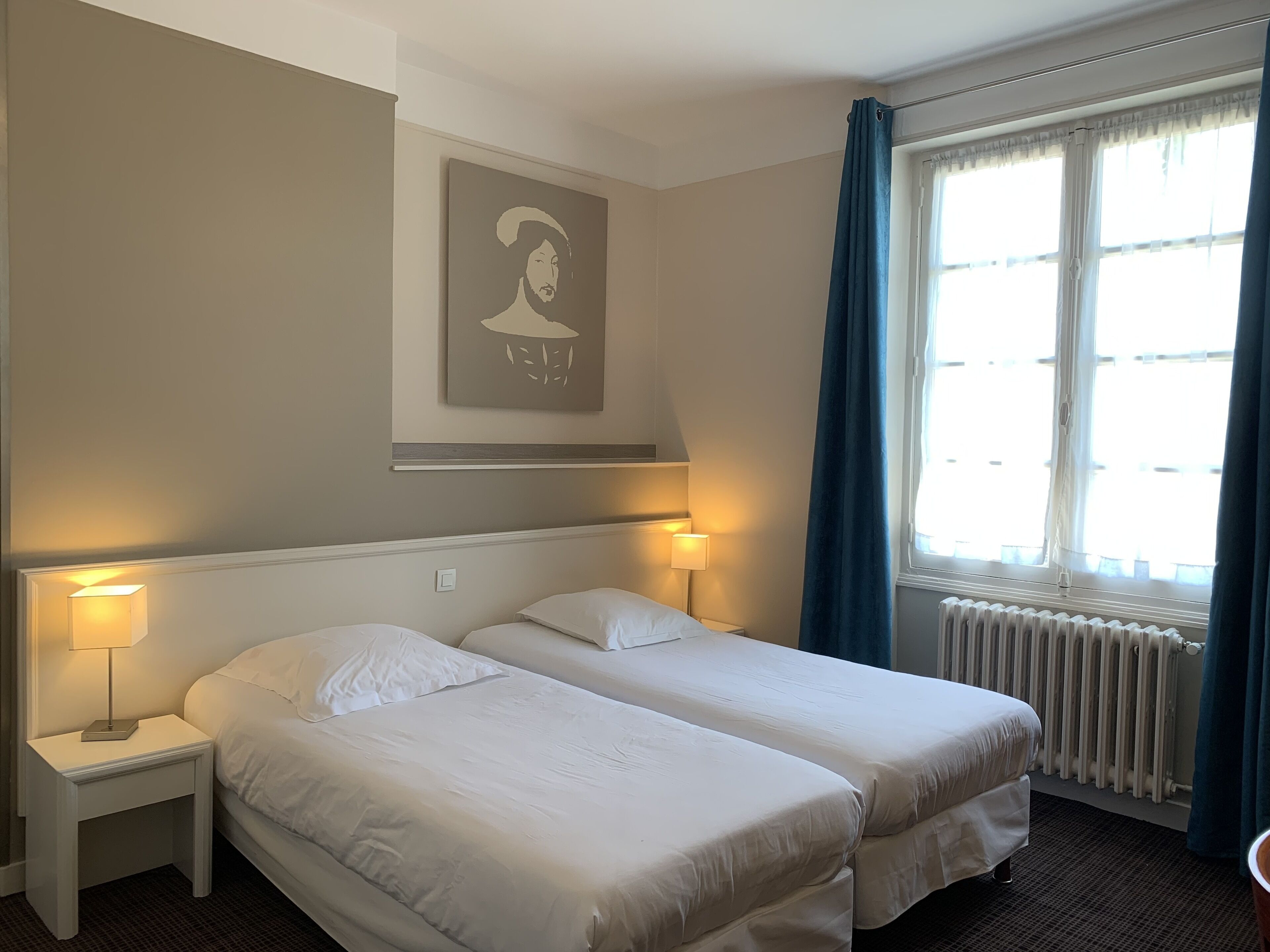 A hotel room with two beds, a radiator, a window with curtains, and a wall-mounted picture.