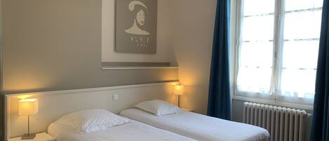 A hotel room with two beds, a radiator, a window with curtains, and a wall-mounted picture.
