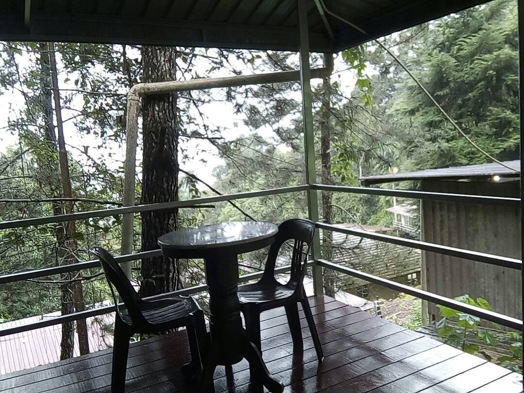 A covered outdoor seating area with a table and chairs, surrounded by a wooden deck and metal railing, overlooking a forested area.
