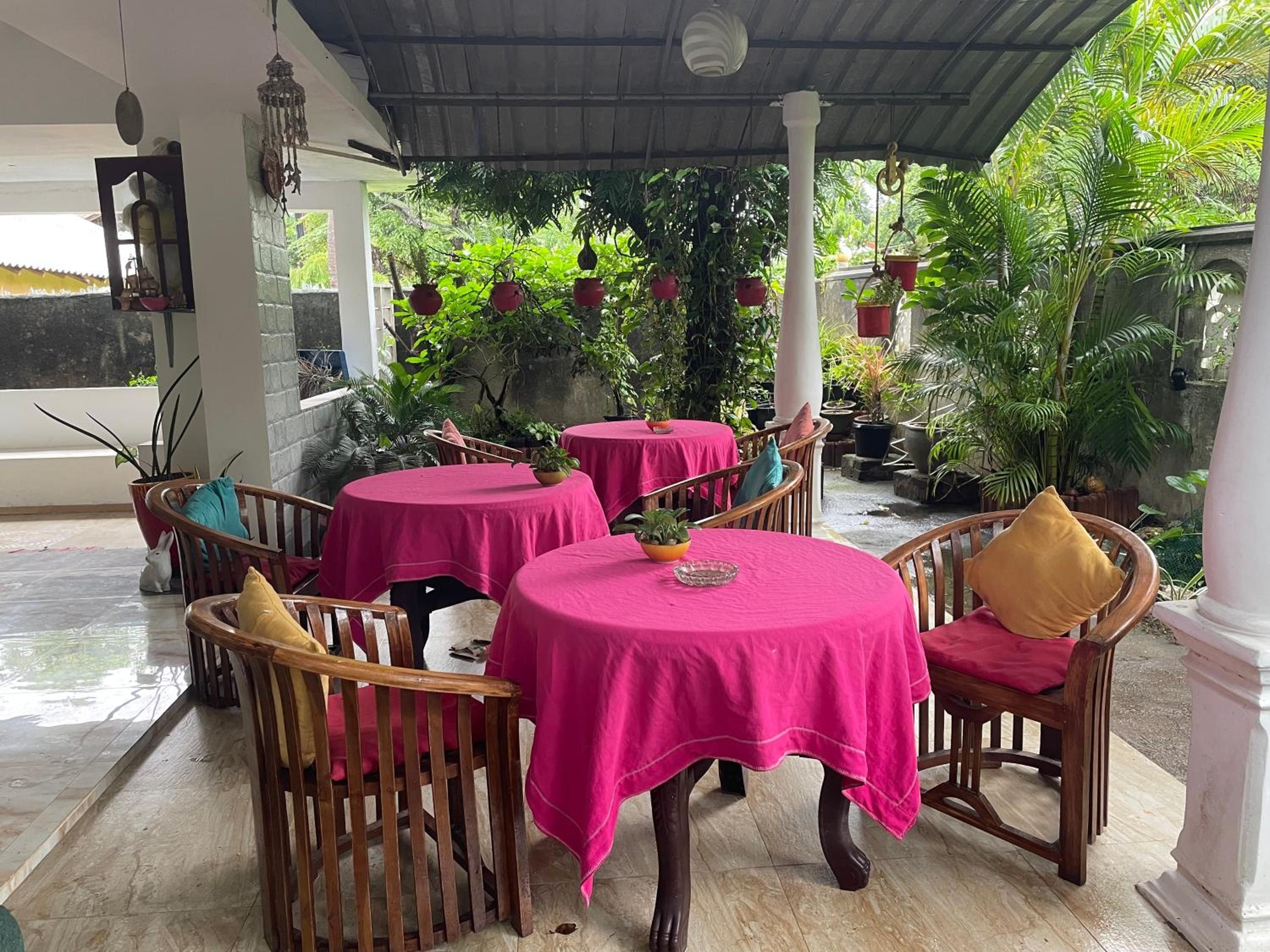 A covered outdoor seating area with round tables and chairs, surrounded by potted plants and greenery.