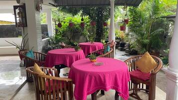 A covered outdoor seating area with round tables and chairs, surrounded by potted plants and greenery.