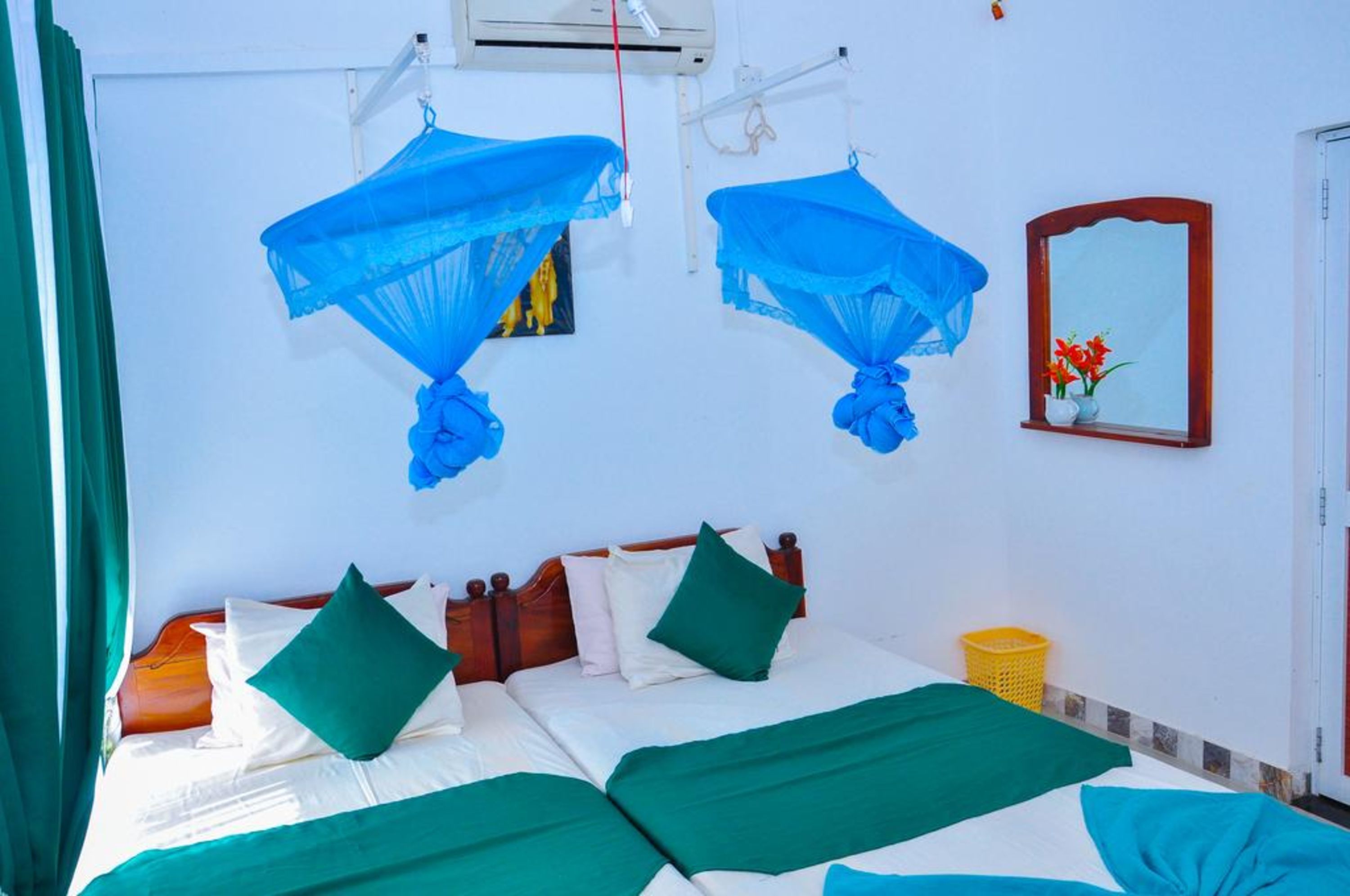 A hotel room with a bed, green bedding, a wooden headboard, a mirror, and a yellow basket.