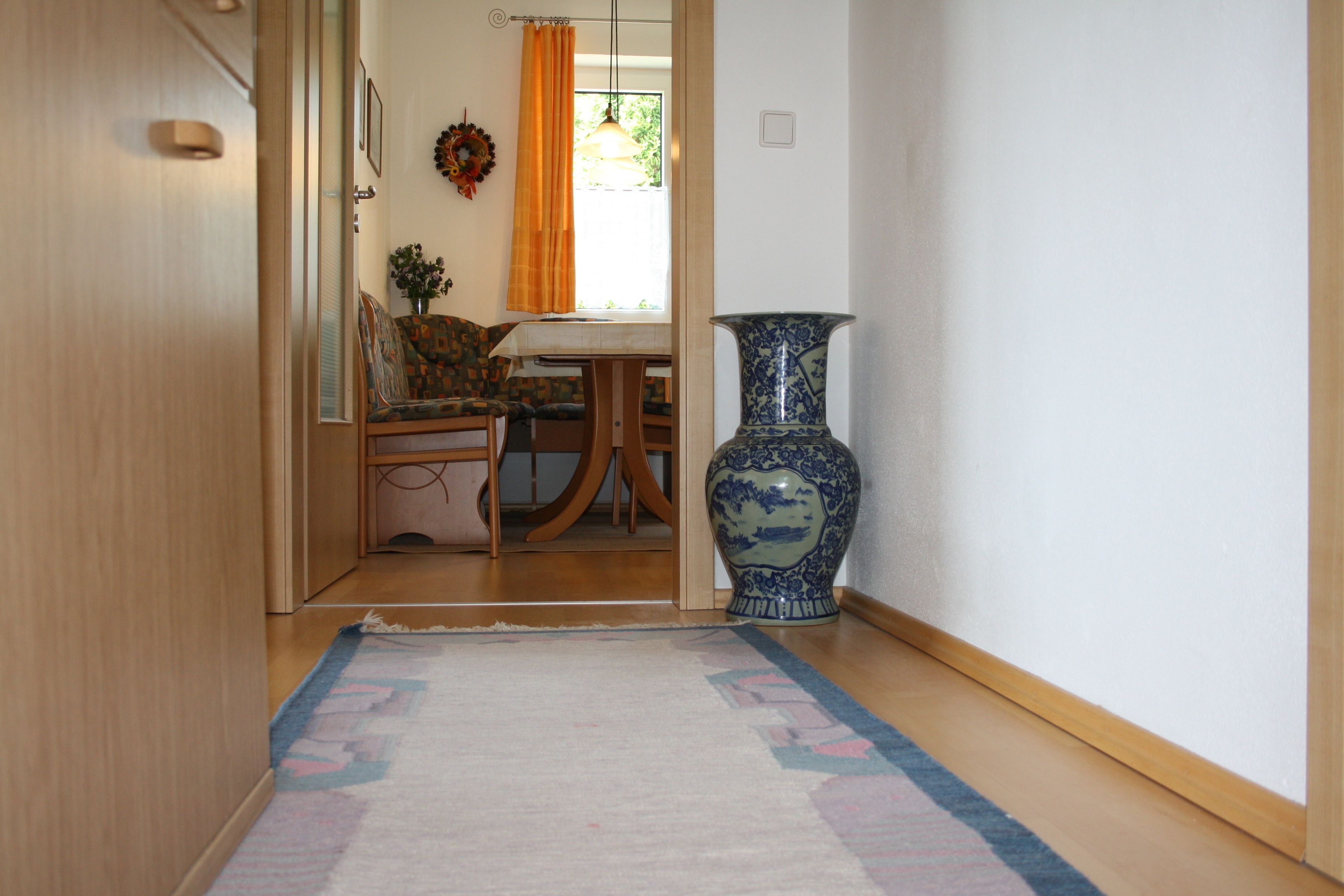A hallway with a patterned rug, a wooden door, a blue and white vase, and a dining area with a table and chairs.