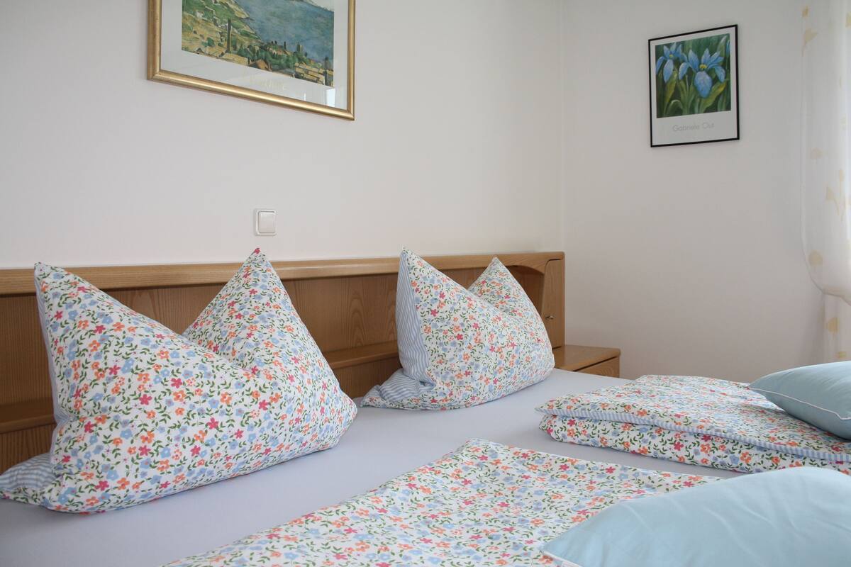 Ein ordentlich bezogenes Bett mit blumigem Bettbezug und Kissen, ein hölzernes Kopfteil und ein gerahmtes Bild an der Wand.