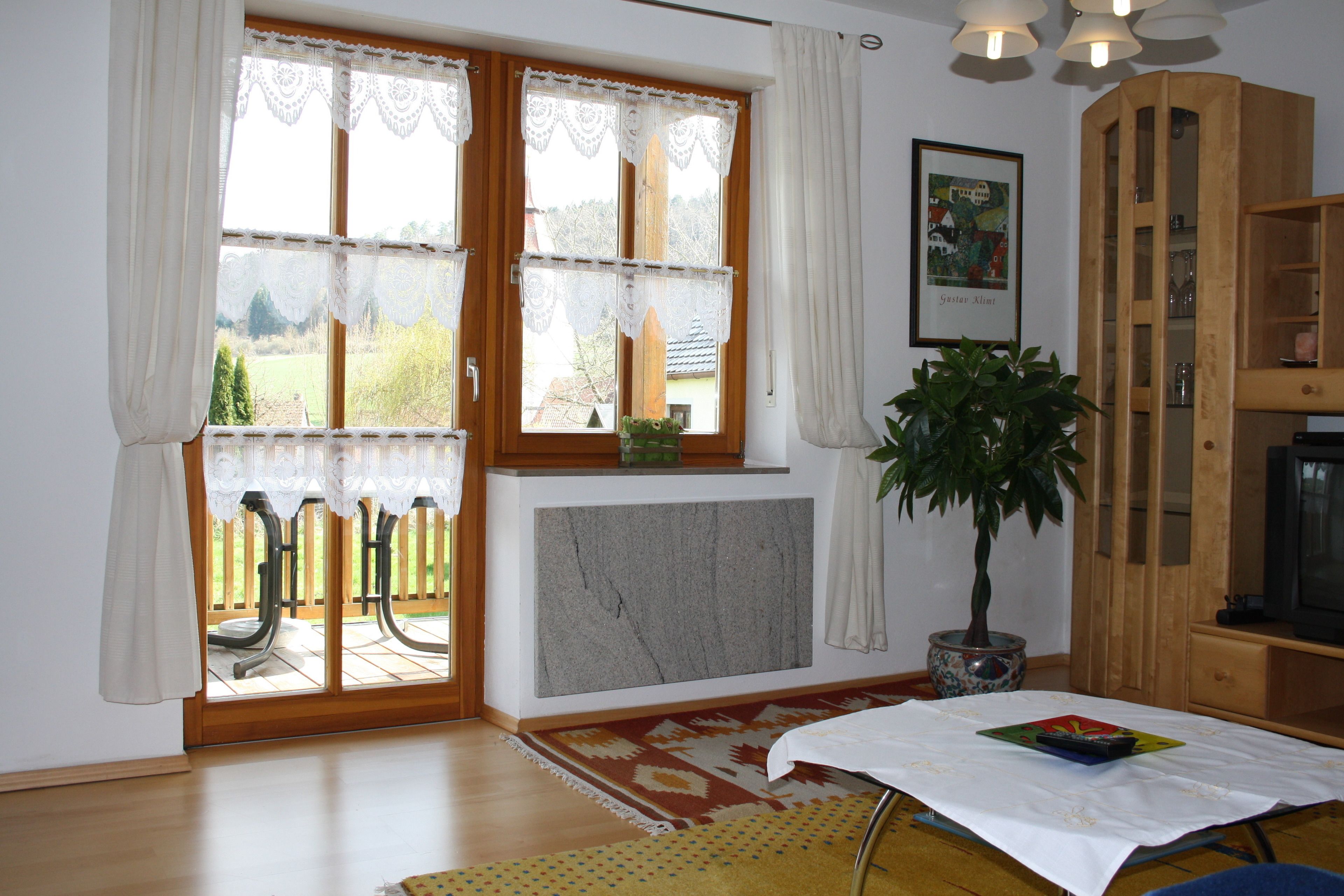 Sala de estar com uma grande janela, uma planta em vaso, uma mesa com toalha e um armário de madeira.