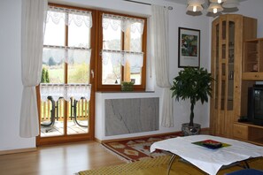 Een woonkamer met een groot raam, een potplant, een tafel met tafelkleed en een houten kast.