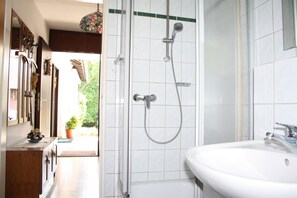 Een badkamer met een douche, wastafel en spiegel. Een deur leidt naar een buitenruimte met een potplant.