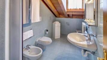 Een badkamer met toilet, bidet en wastafel, betegeld in lichtblauw, met een houten plafond en een raam.