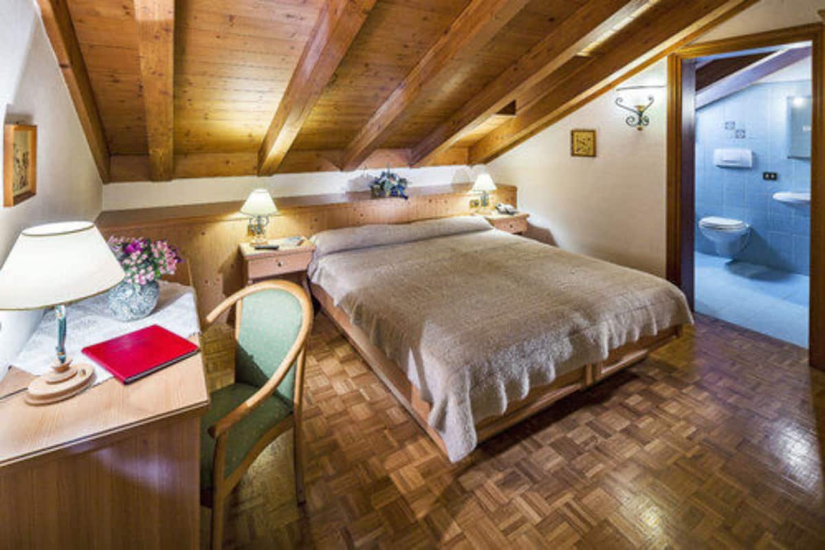 A cozy bedroom with a wooden ceiling, a bed with a grey blanket, a nightstand with a lamp, a chair, and a view of a bathroom with a toilet and sink.