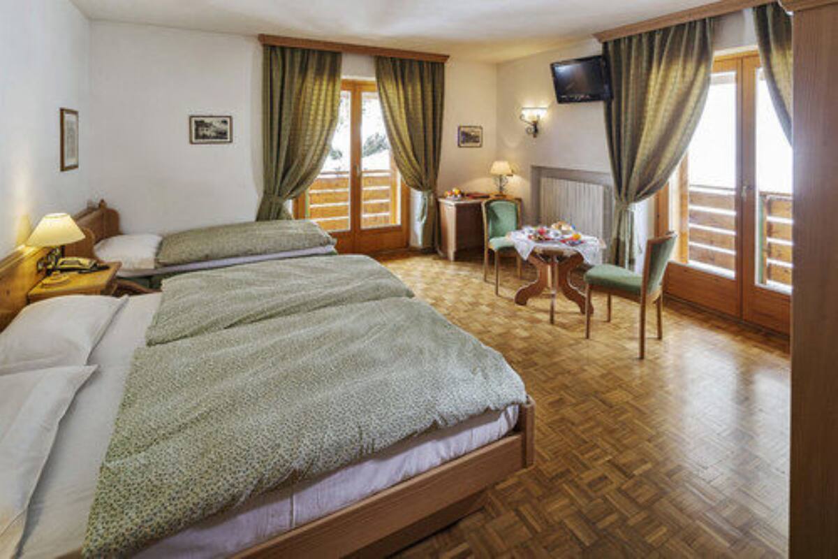 A hotel room with two single beds, a small table with a chair, a TV mounted on the wall, and a window with curtains.